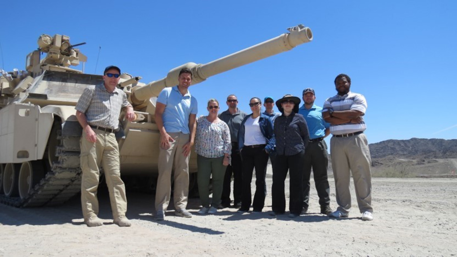 Nine people stand in front of an Abrams tank on a sunny day in the desert.