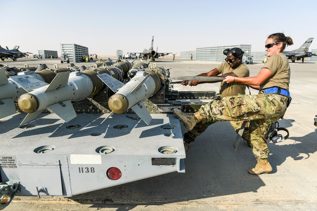 Two airman pull cords over a pallet containing bombs.