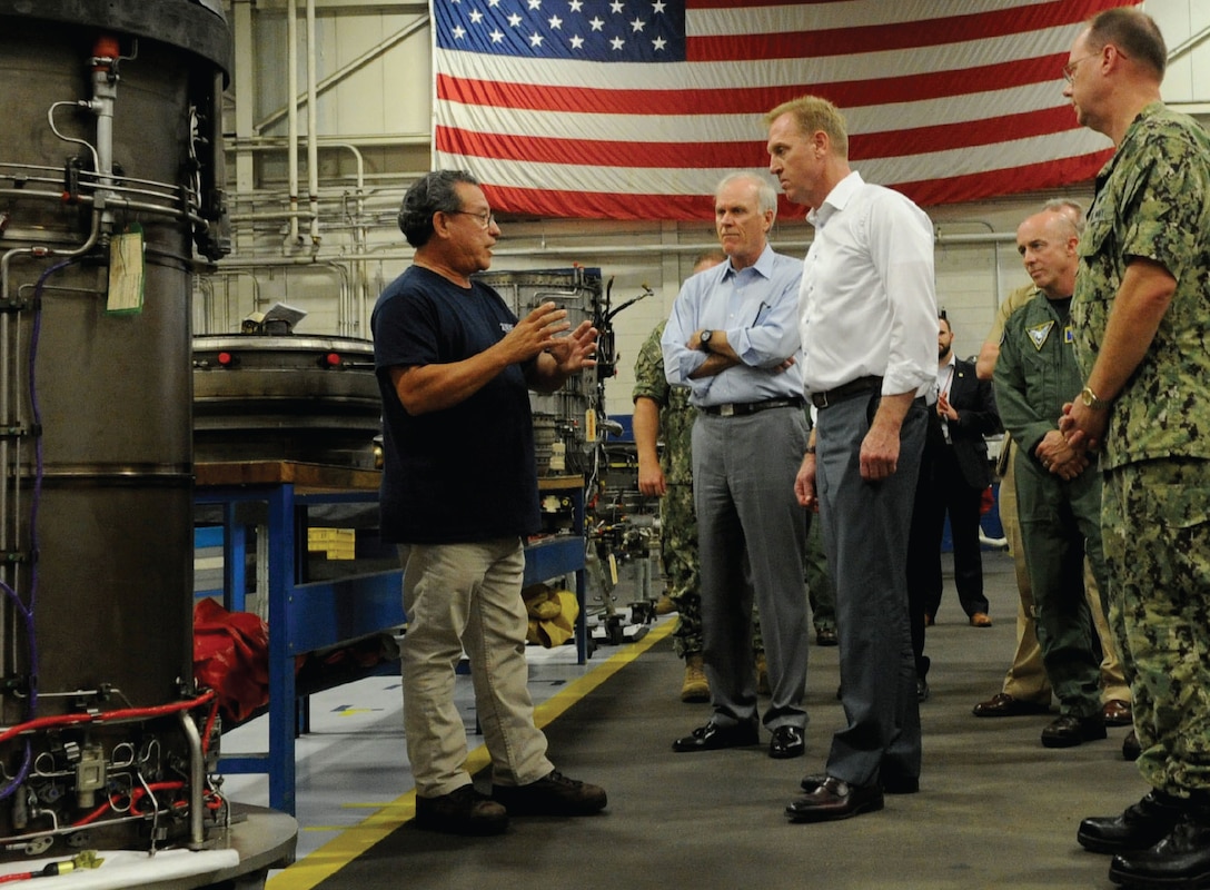 Deputy Defense Secretary Patrick M. Shanahan talks to people in utility room with an American flag on a wall.