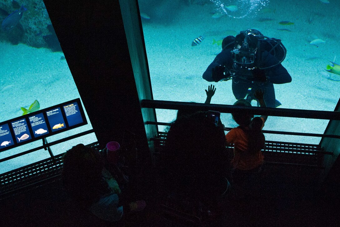 A diver inside an aquarium tank interacts with children on the other side.