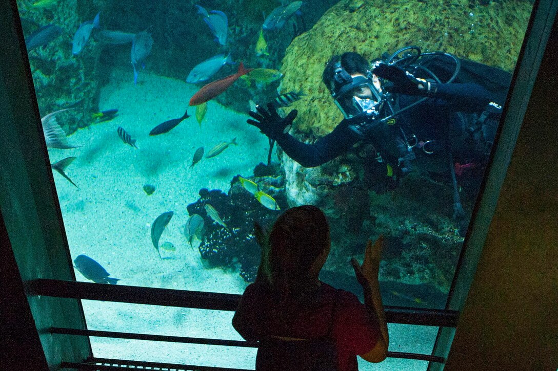 A diver inside an aquarium tank interacts with children on the other side.