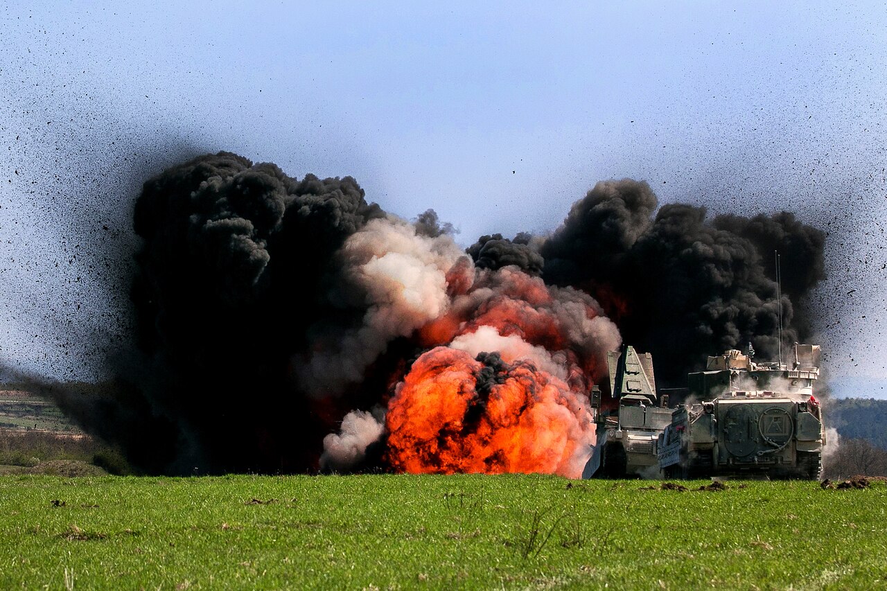 Black clouds of smoke flank a fireball from a detonation in a field.