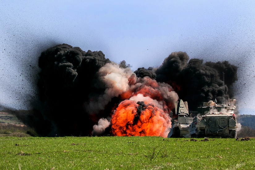 Black clouds of smoke flank a fireball from a detonation in a field.