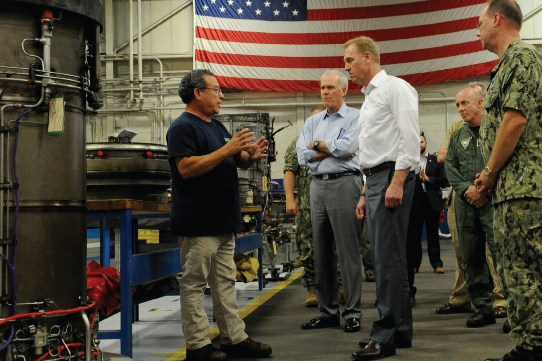 Deputy Defense Secretary Patrick M. Shanahan talks to people in utility room with an American flag on a wall.