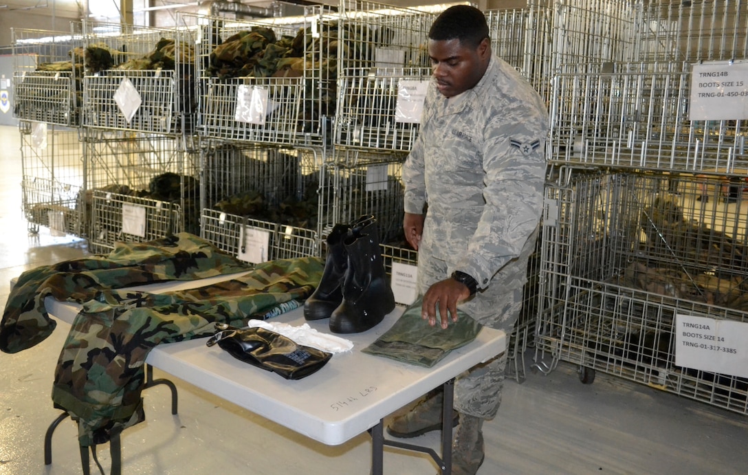 An airman displays military clothing on a table in front of bins of other military clothing