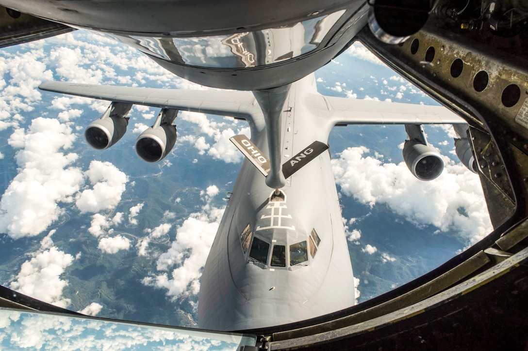 The boom of one aircraft connects to another aircraft flying in a cloud-speckled sky.