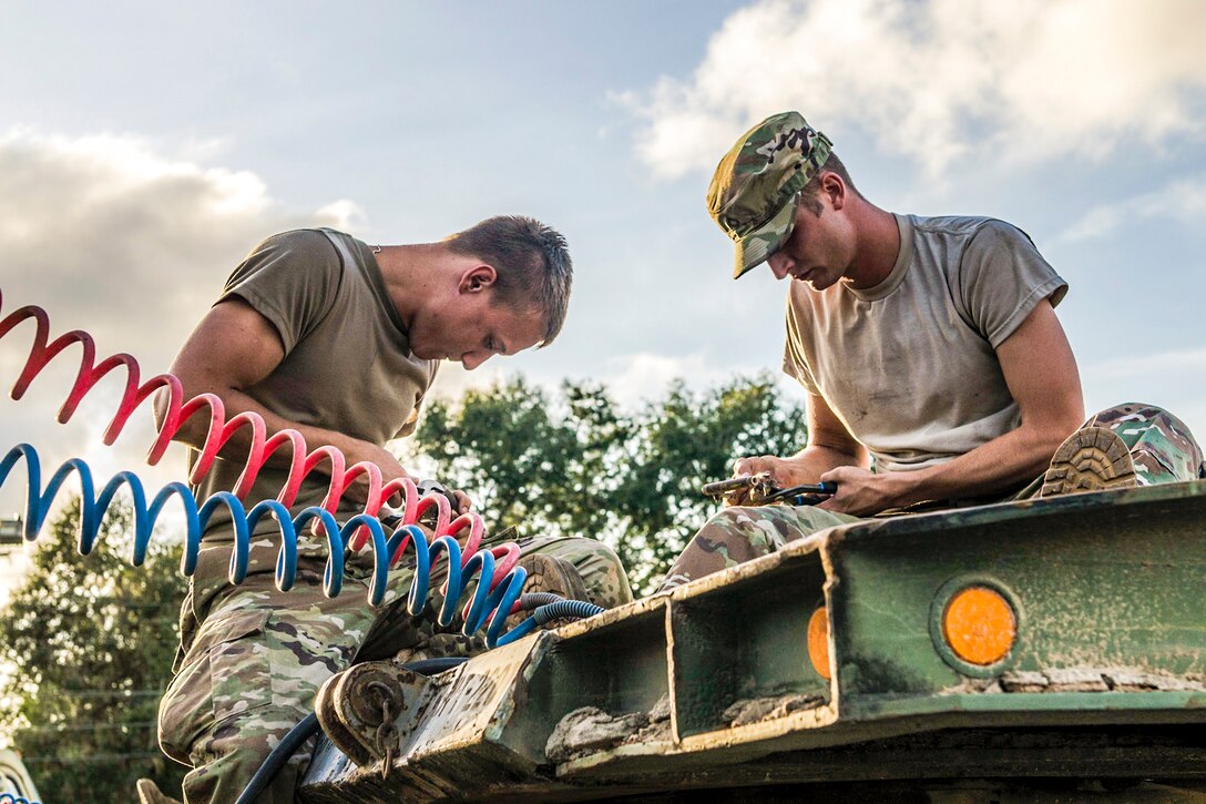 Two soldiers sit atop a green piece of equipment and look down at their work.