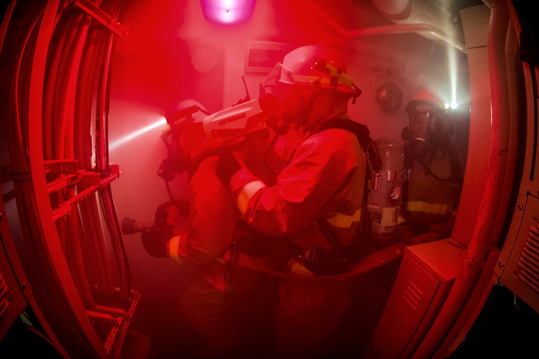 Three firefighters move through smoke wearing protective clothing on a Navy vessel