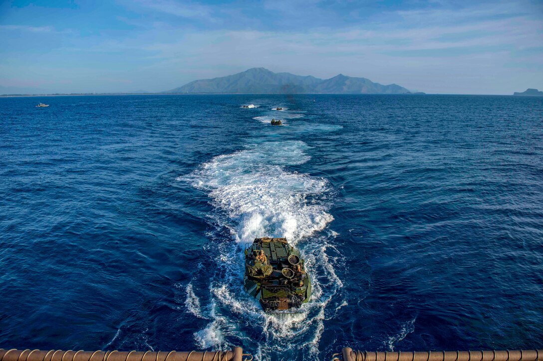 Amphibious military vehicles make their way toward a ship in the sea.