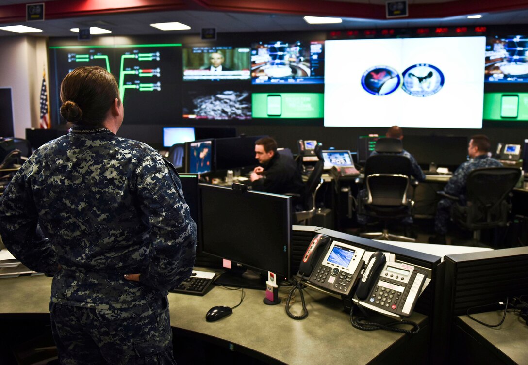 A sailor stands in an operations center-type room with a wall of screens and sailors seated at banks of computers.