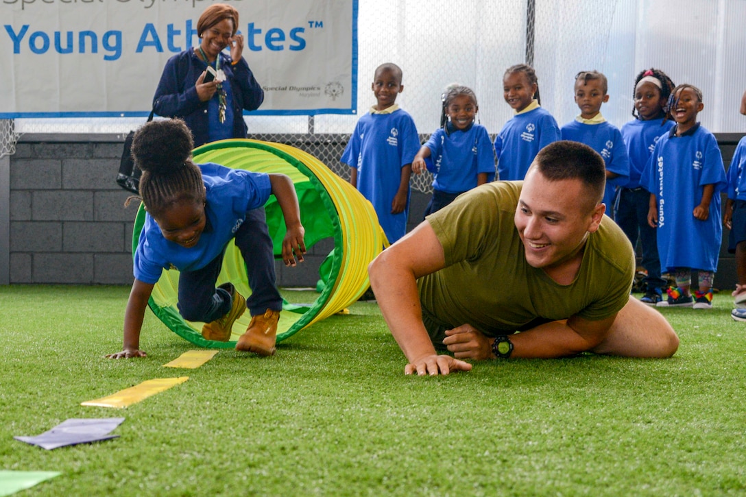 A Marine crawls on grass next to a little girl emerging from an obstacle as a line of children and one adult watch smiling.