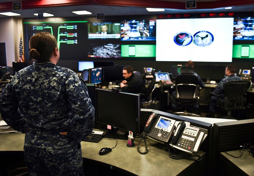 A sailor stands in an operations center-type room with a wall of screens and sailors seated at banks of computers.