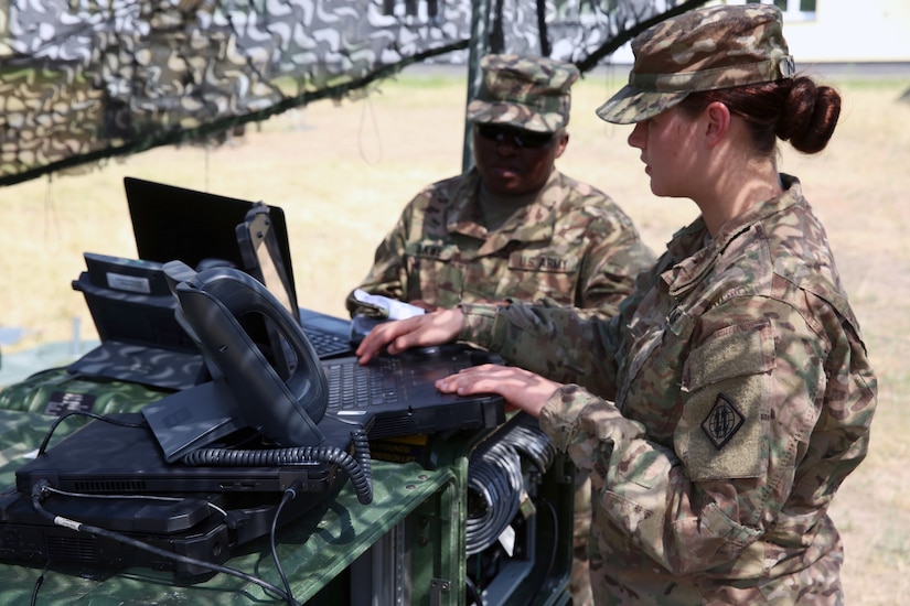 Two soldiers work on computers on a table outside in desert-type terrain.