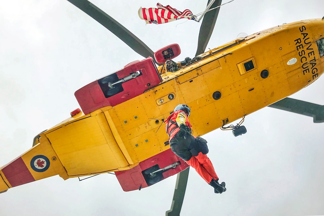 A Coast Guardsman, shown from below, hangs out of a yellow helicopter in flight.