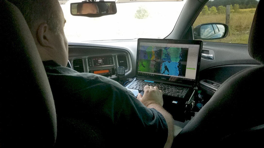A man is in a car looking at a laptop displaying storm radar photos.