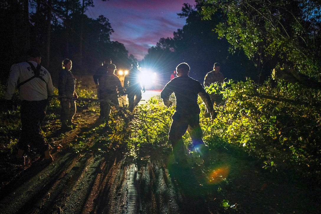 Several guardsmen clear brush and downed trees from a road at night.