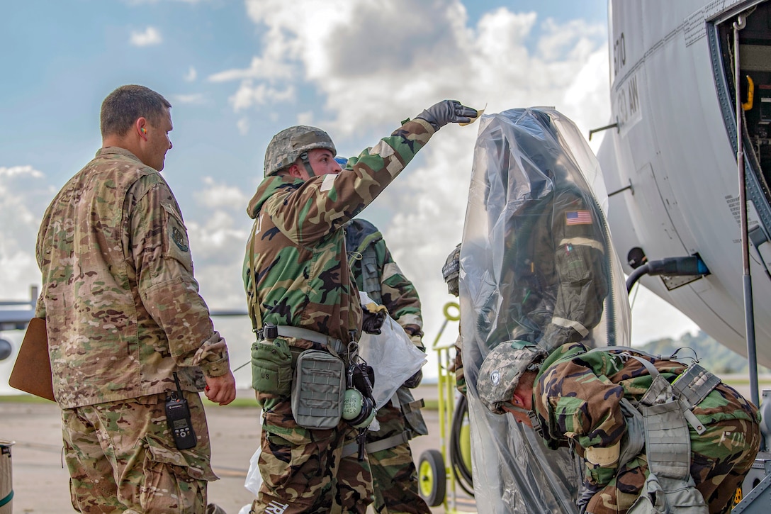 Airmen stand outside an airplane and place a person-sized plastic bag over another airman.