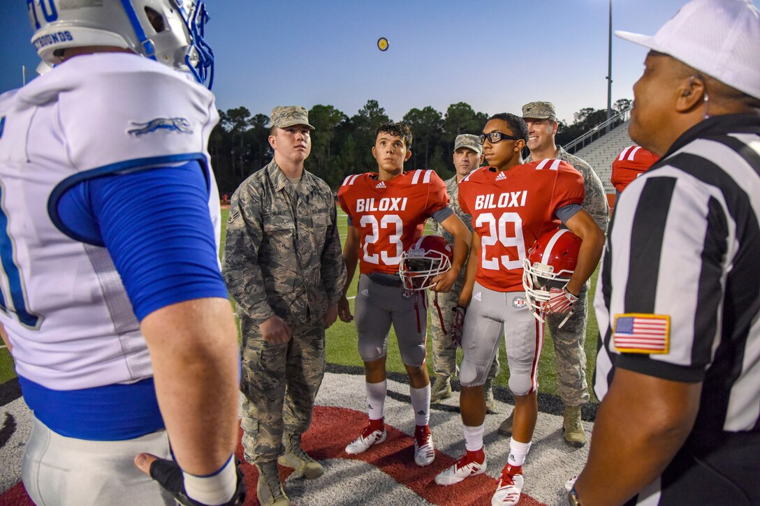 High school football players, a referee, and an airmen watch a coin flip through the air before a football game.