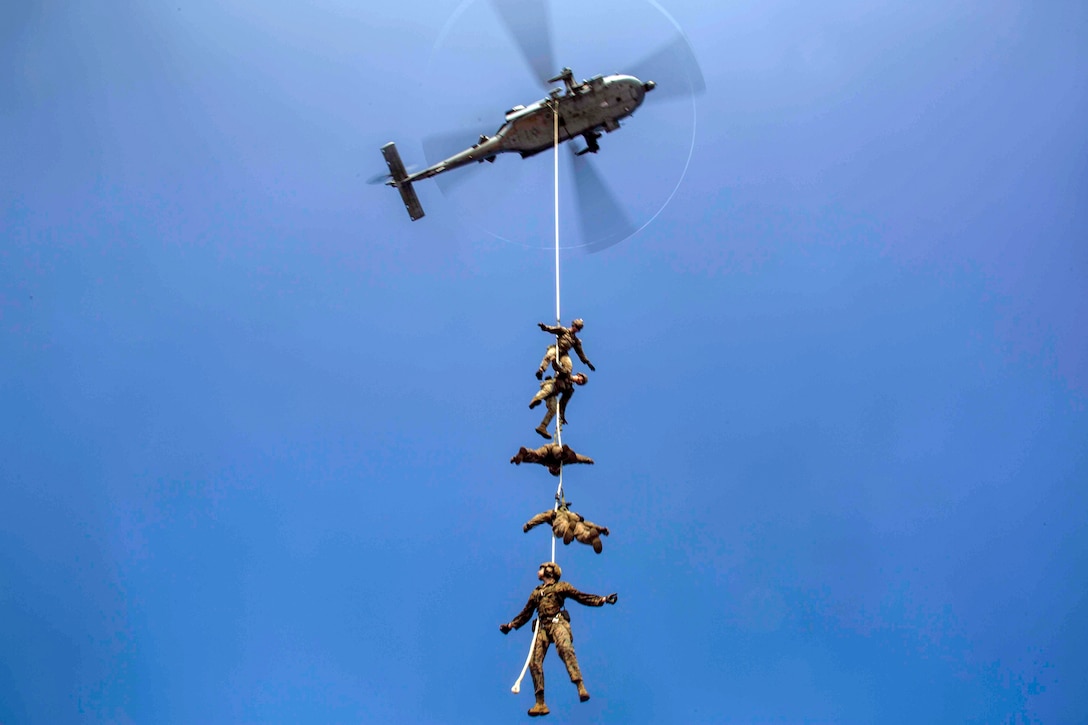 Five Marines, bound to a rope, hang midair, carried by a helicopter.