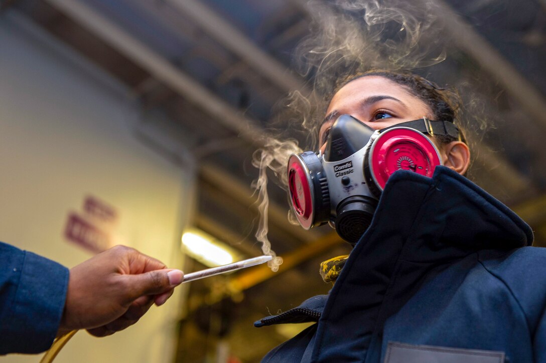 A sailor wears a breathing mask while another sailor waves a burning stick near the mask.