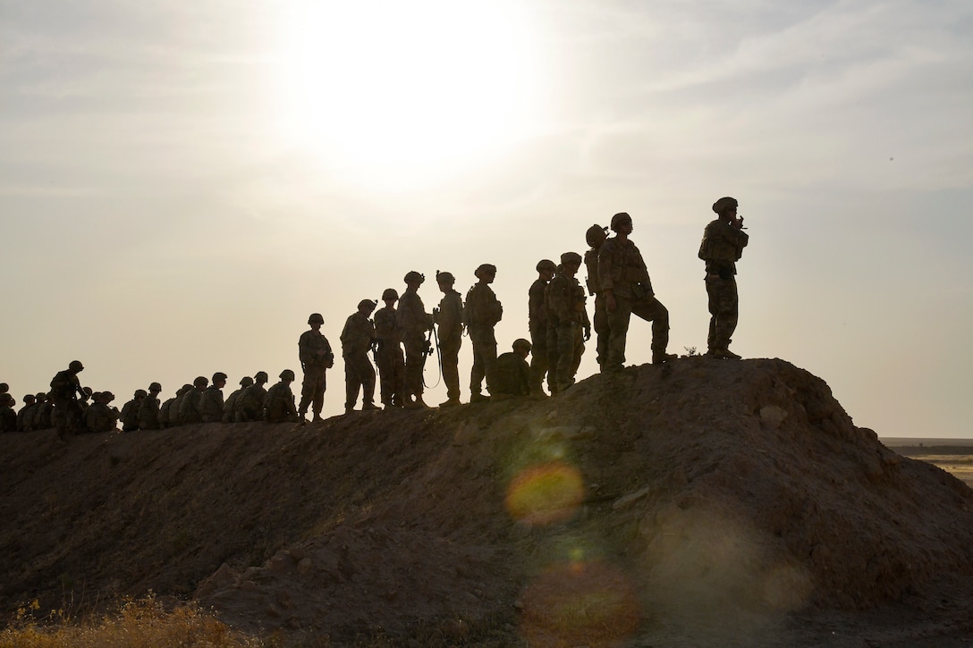 More than a dozen soldiers, shown in silhouette, stand and look out on a hill.