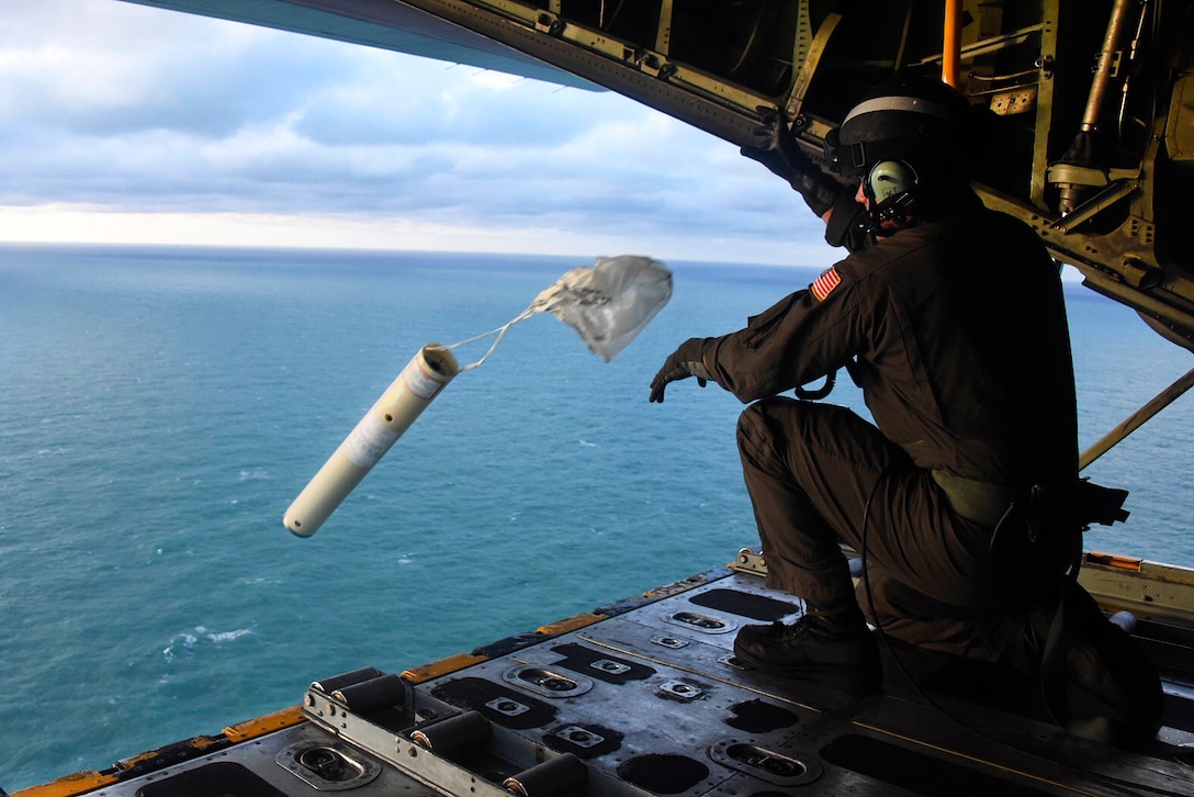 A Coast Guardsman tosses a cylindrical object out of the back of an open aircraft.