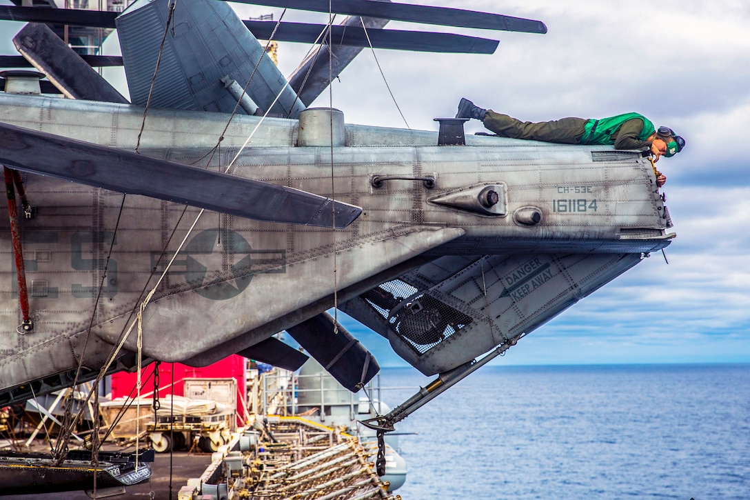 A Marine lays on top of the end of an aircraft clearing rust, over the open ocean.