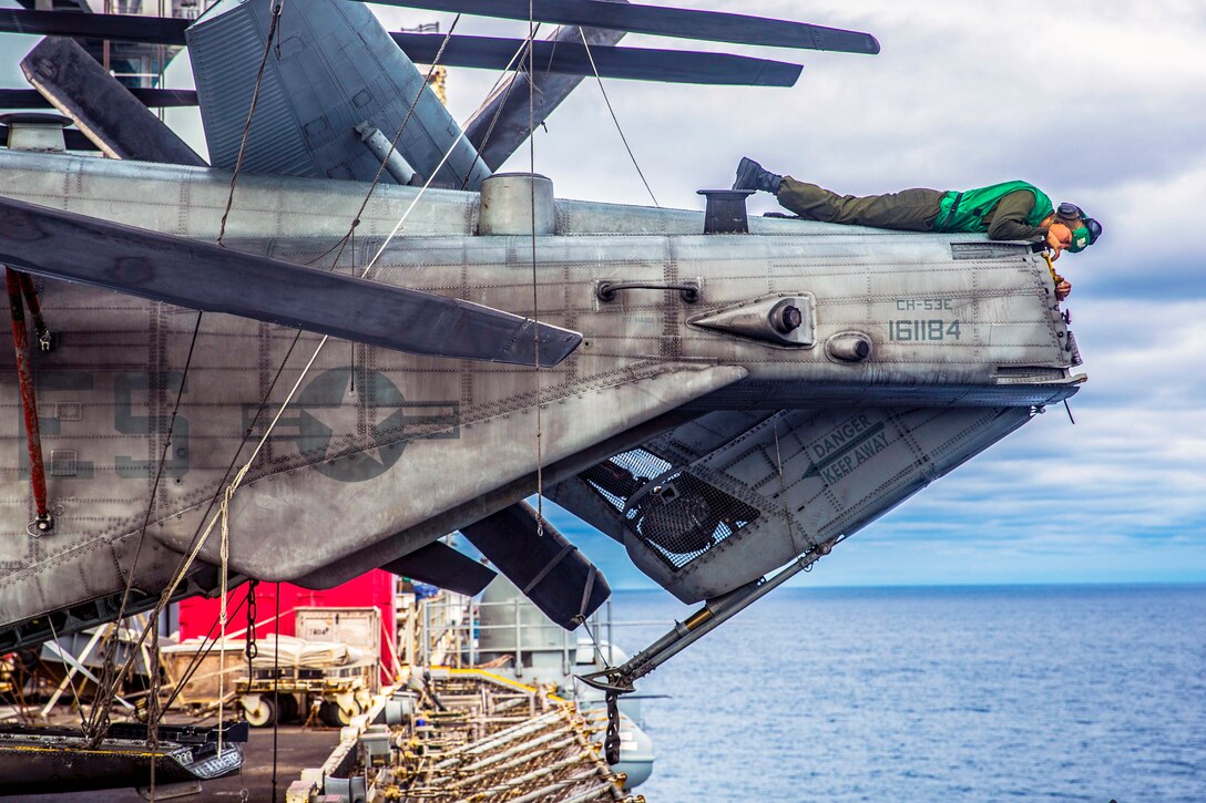 A Marine lays on top of the end of an aircraft clearing rust, over the open ocean.