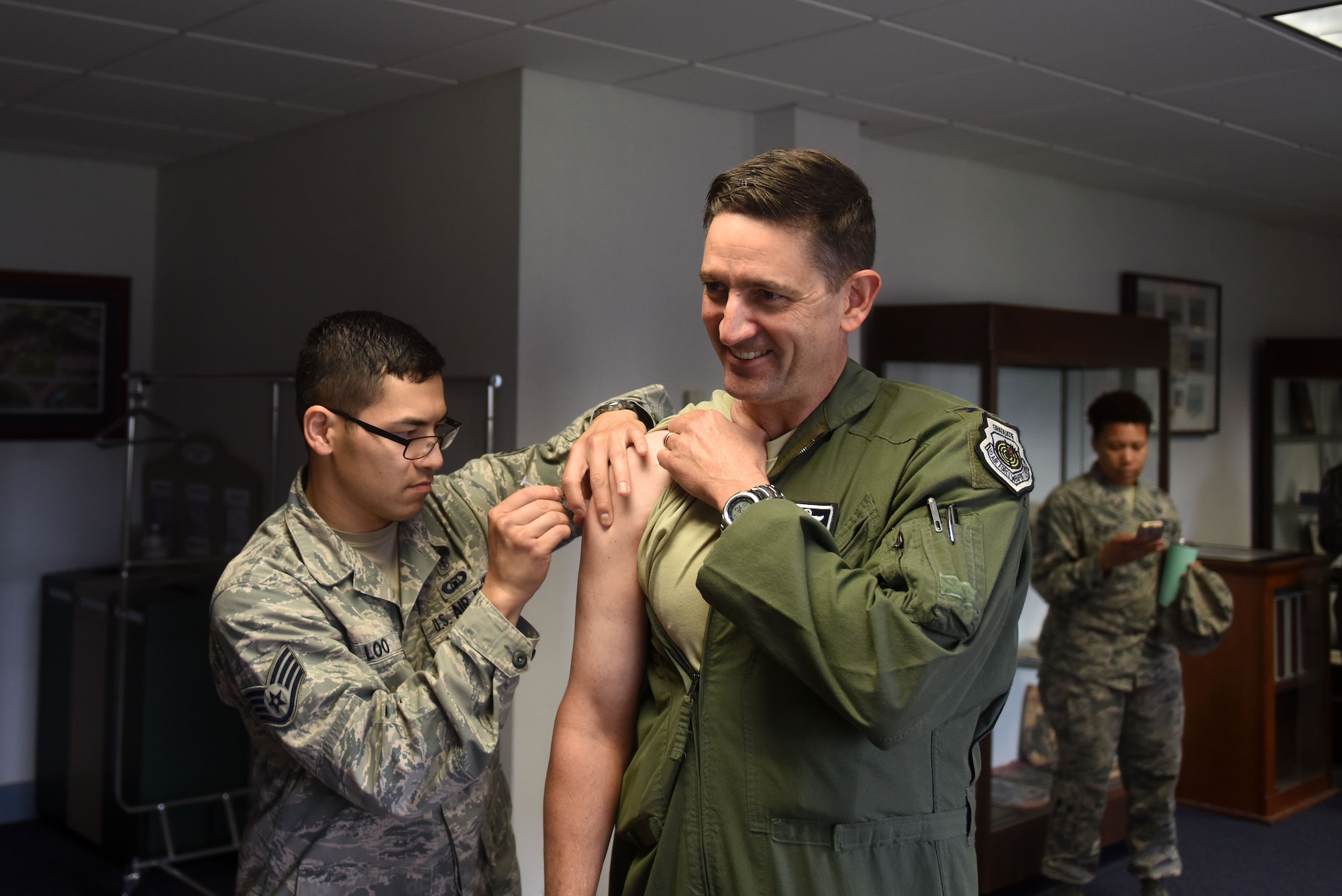 A man with dark hair wearing a flight suit receives a flu shot from a man with dark hair and glasses wearing the Airman Battle Uniform.