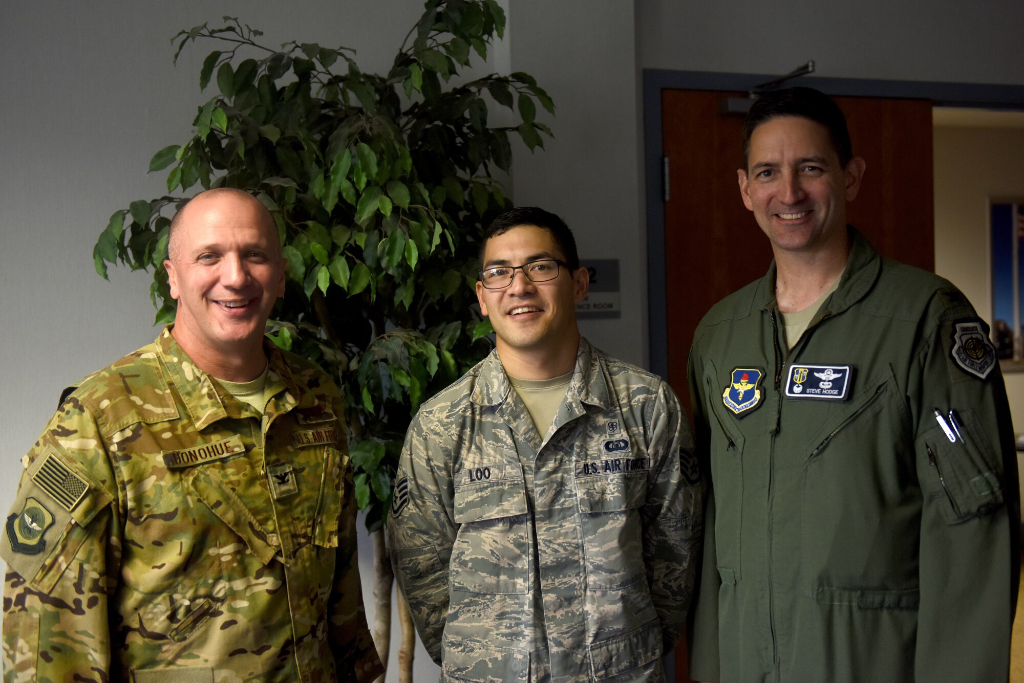 Three men stand smiling at the camera. The farthest left is smiling and wearing the Operational Camouflage Patter uniform, the middle is a man with dark hair and glasses wearing the Airman Battle Uniform and the far right man is wearing a green flight suit.