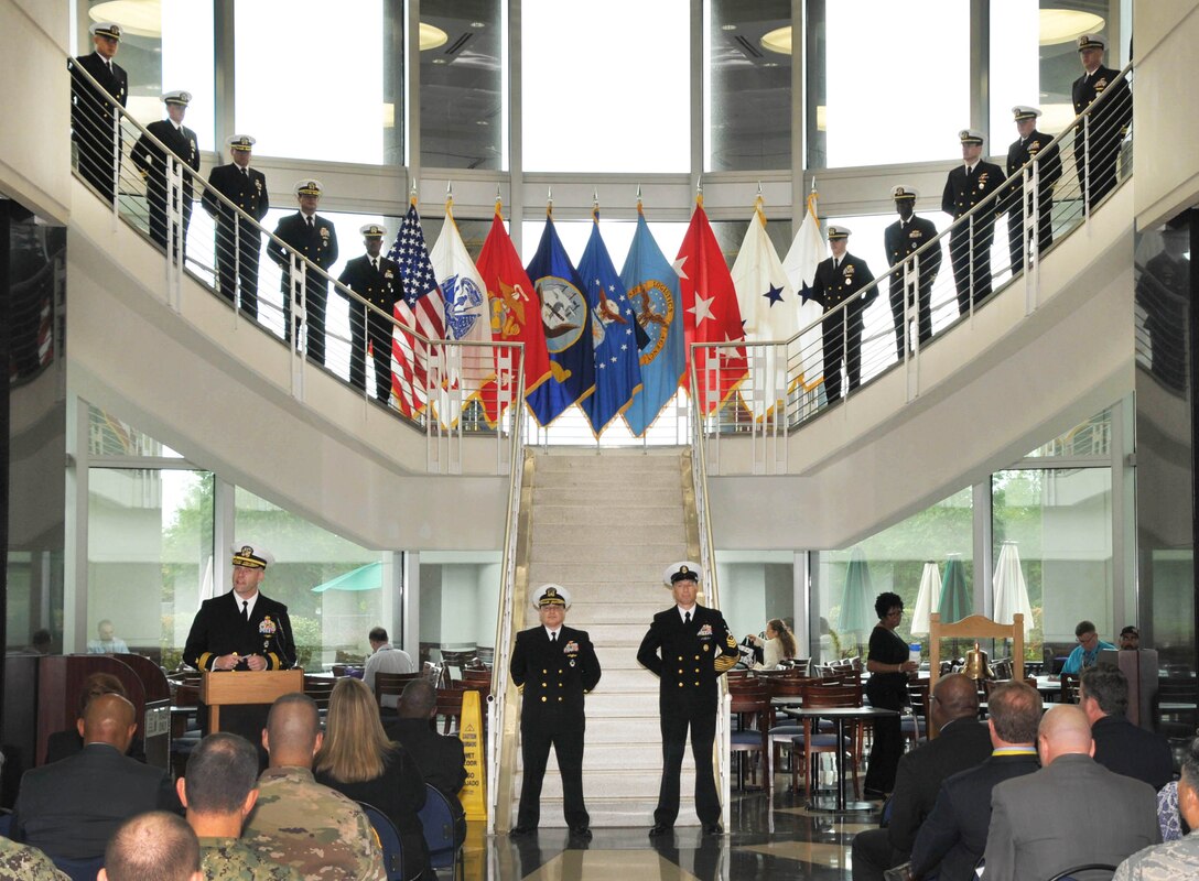 DLA Land and Maritime Commander Rear Admiral John Palmer, left, addresses the audience as officers and sailors stand in formation along the staircase in background.