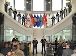 DLA Land and Maritime Commander Rear Admiral John Palmer, left, addresses the audience as officers and sailors stand in formation along the staircase in background.