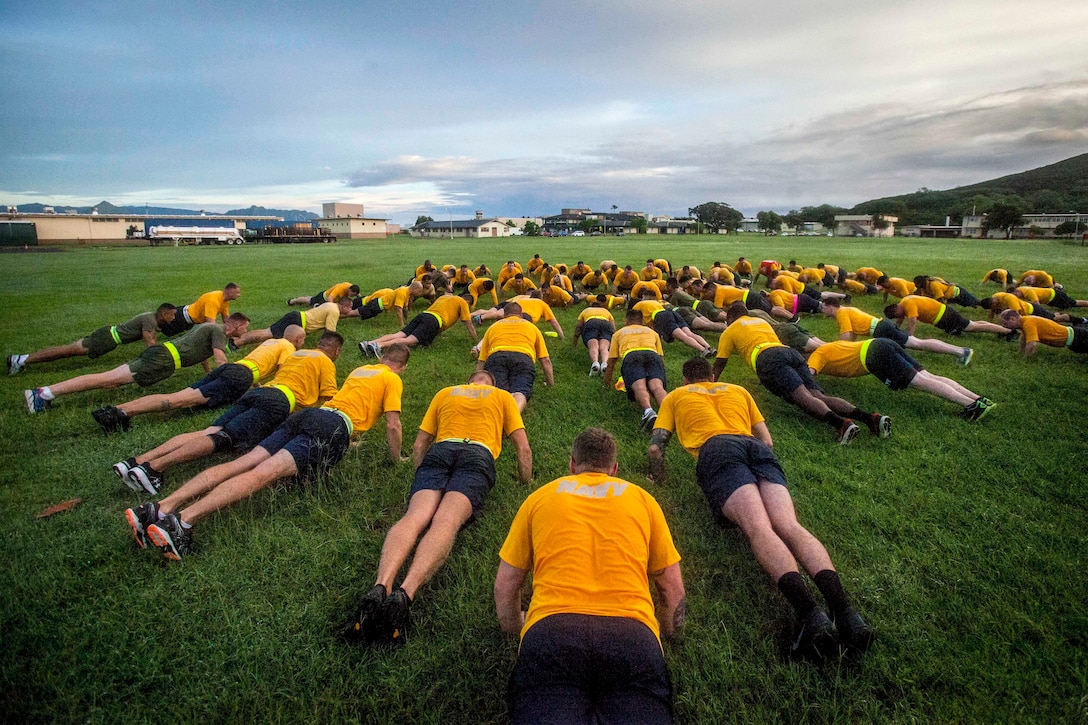 Service members in yellow shirts and blue shorts do pushups in a circle on a grass field.