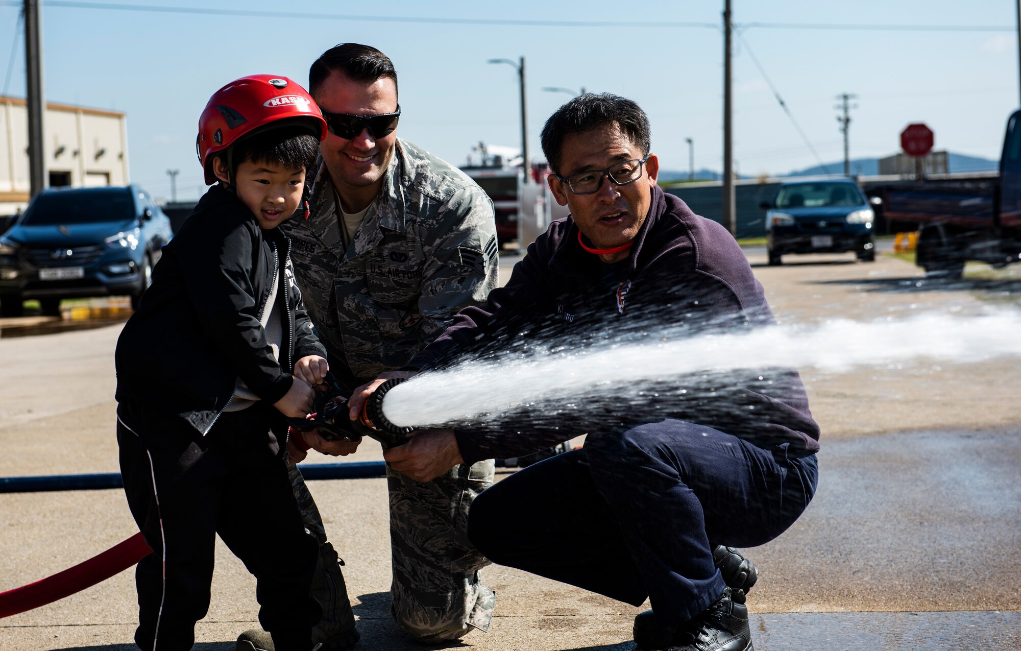 U.S. Air Force Senior Airman Anthony Pipes (middle) and Ki Song Kim (right), 8th Civil Engineering Squadron fire fighters, help a student use a fire hose during an open house at Kunsan Air Base, Republic of Korea, Oct. 11, 2018. The Kunsan Fire Department welcomed 60 students from Su Song Elementary School to show them some of the equipment they use and teach them about fire safety. (U.S. Air Force photo by Senior Airman Stefan Alvarez)