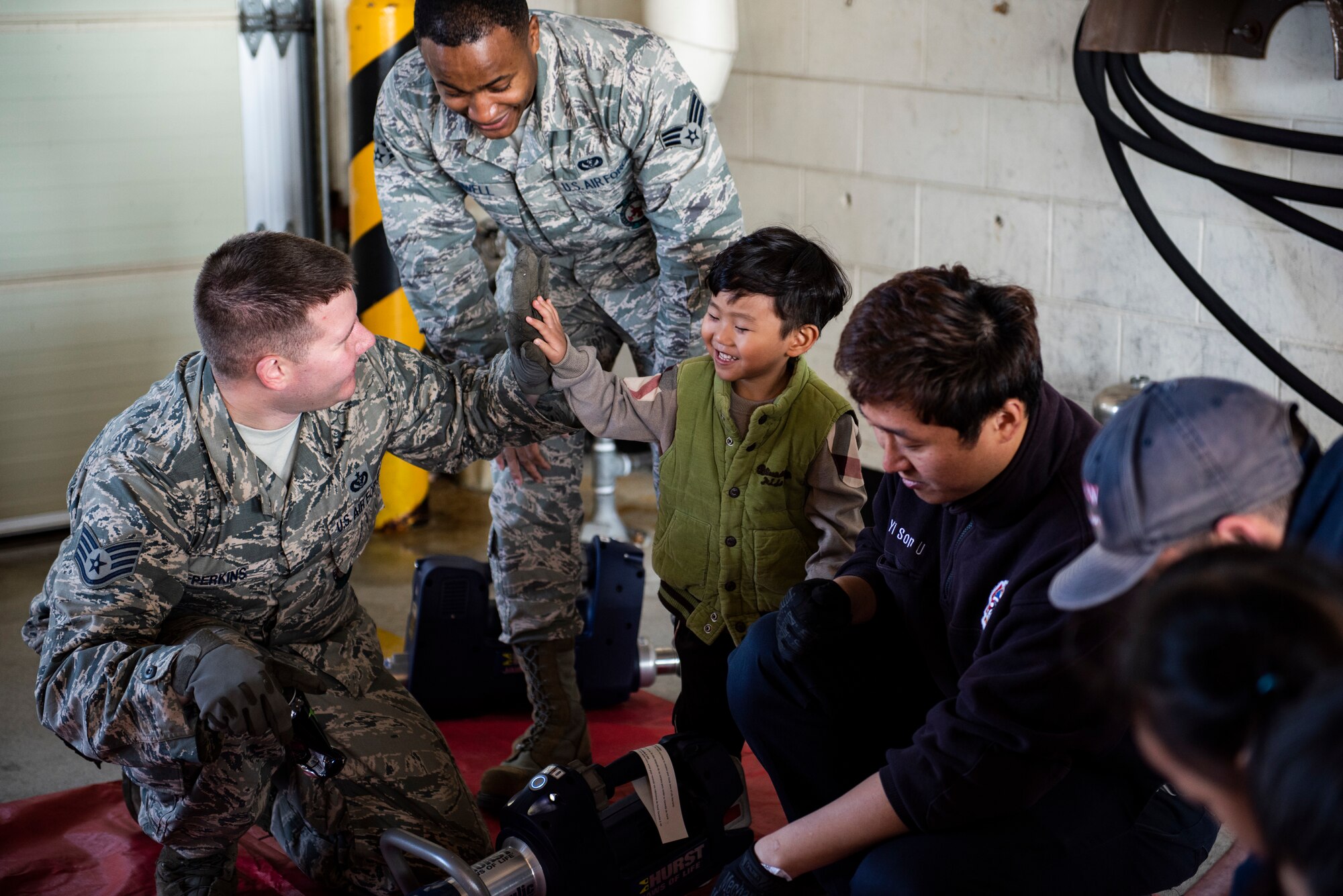 U.S. Air  Force Staff Sgt. Jonathan Perkins (left) and Senior Airman Michael Rowell (middle), 8th Civil Engineering Squadron fire fighters, help a student Su Song Elementary use a metal cutting tool during an open house at Kunsan Air Base, Republic of Korea, Oct. 11, 2018. Tools like this are often needed if fire fighters need to get through large metal objects or gain access to locked doors. (U.S. Air Force photo by Senior Airman Stefan Alvarez)
