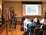 Man stands before a group of seated people in a large room. An image is projected on a screen.