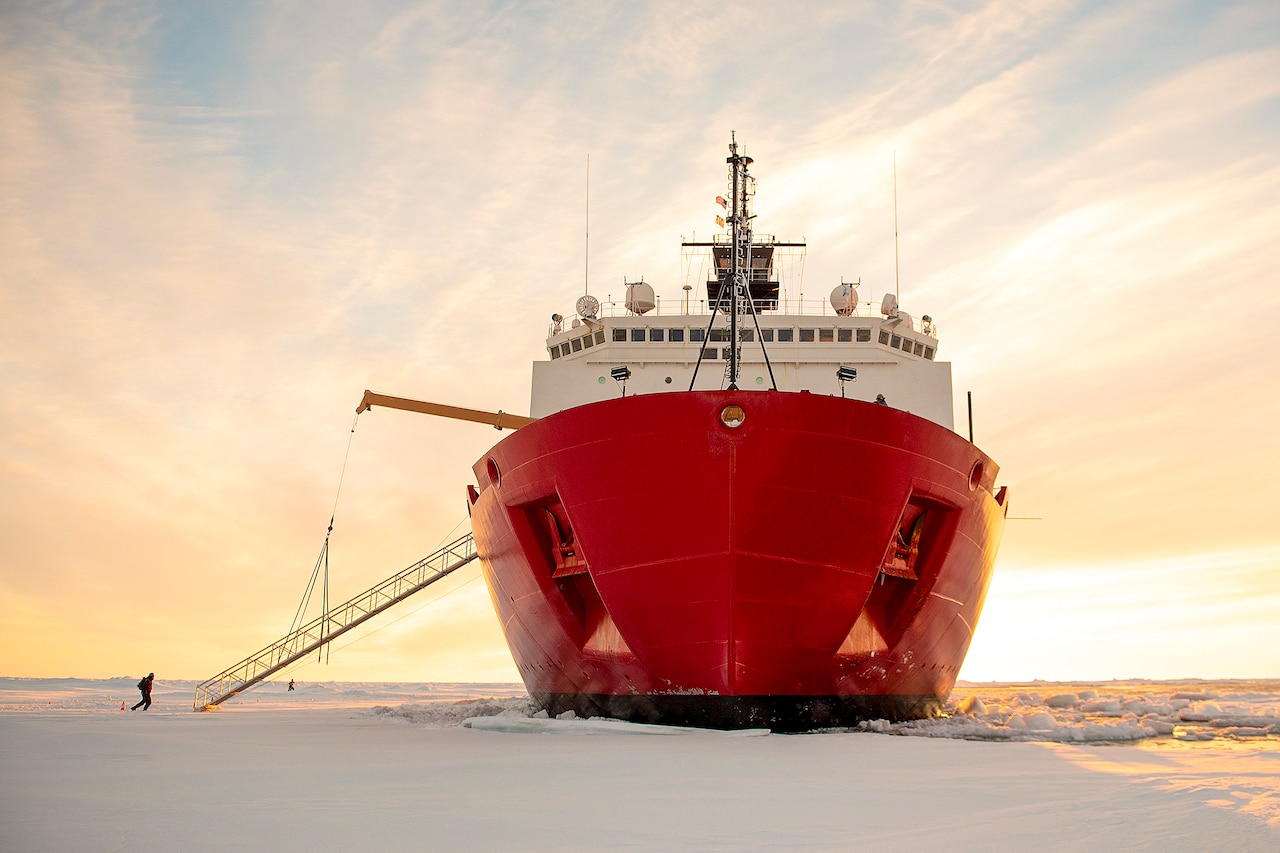 A person approaches a giant red ship sitting on an ice field against a pink-streaked sky.