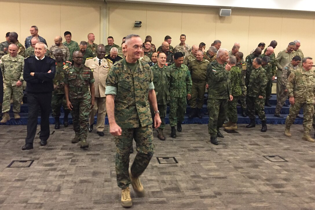 Marine Corps Gen. Joe Dunford stands in front of a group of multinational defense leaders standing on blue risers in a room.