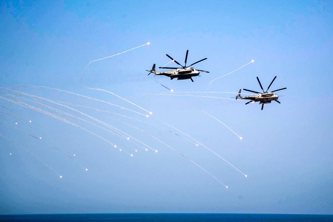 Two helicopters fly between flares over open water.