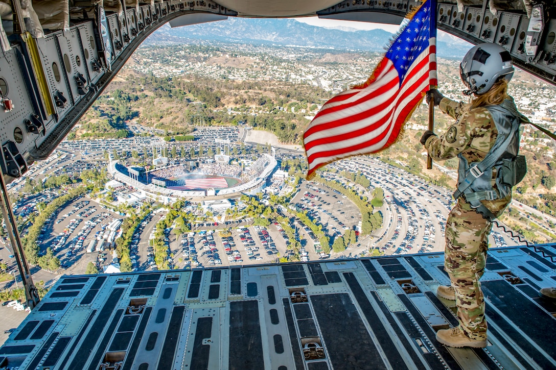 An Airman flies the American Flag out of the back of an airplane flying over a baseball stadium.