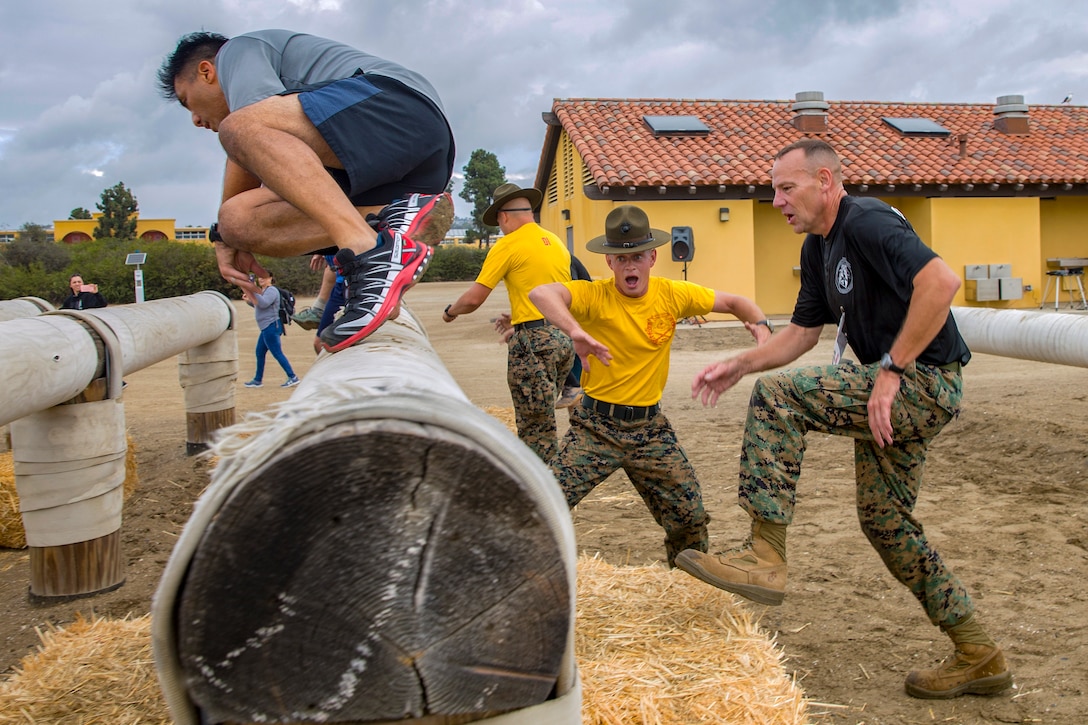 Two participants climb log and hay obstacles while being encouraged by Marine Corps Drill Sergeants.