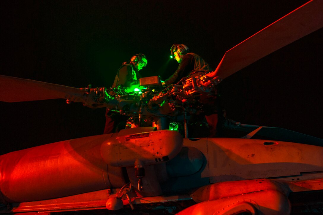 Two sailors work atop a helicopter against a dark sky, illuminated by red and green light.