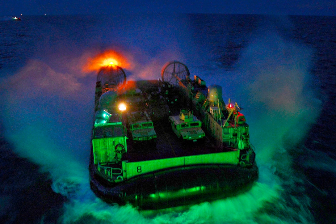 A landing craft, air cushion approaches splashes in water at night.