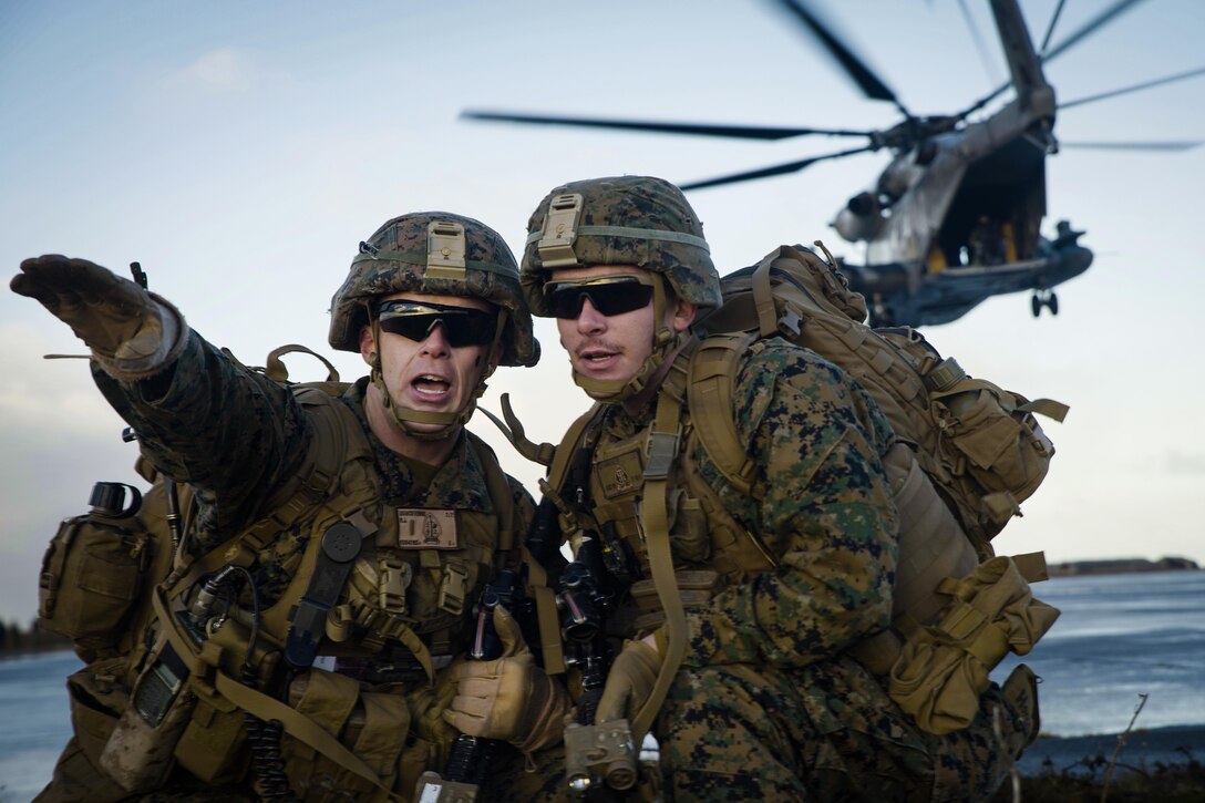 A Marine gestures while giving directions to a team member as a helicopter flies away over water.
