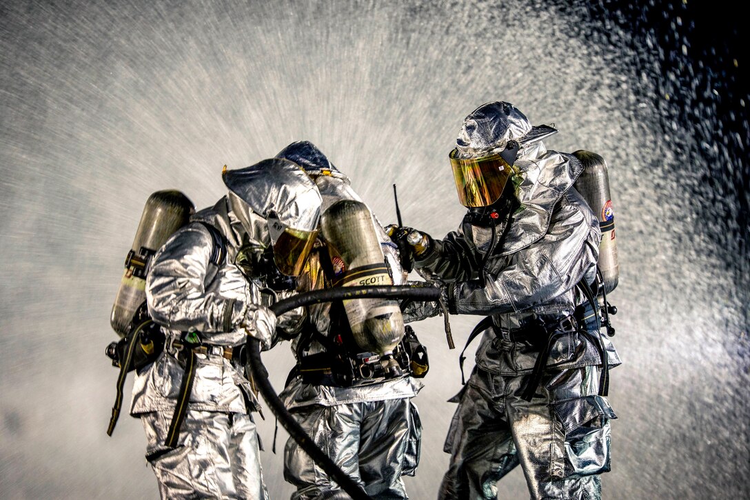 Three firefighters in silver protective gear hold a hose spraying water, which fills the frame.