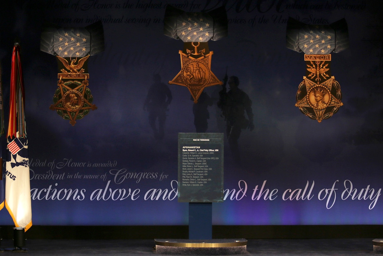 A plaque sits in front of a blue wall with three large recreations of Medals of Honor displayed on it.