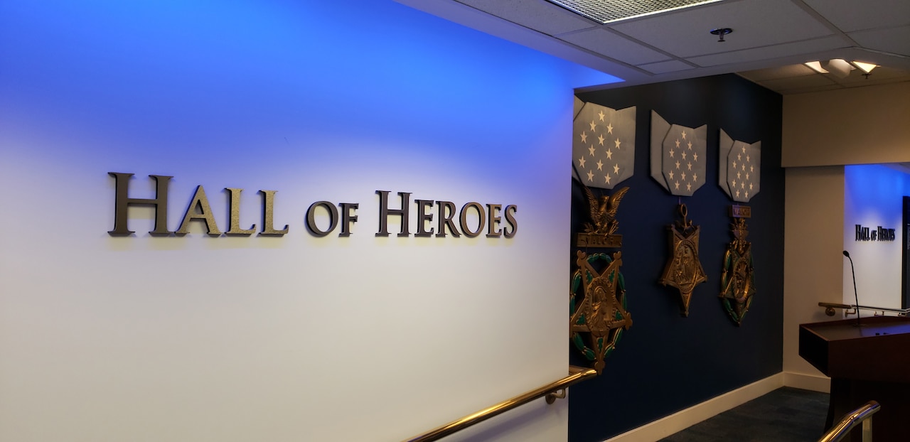 "Hall of Heroes" is posted in gold letters on a white wall, with a blue wall  with large medals displayed on it in the background.