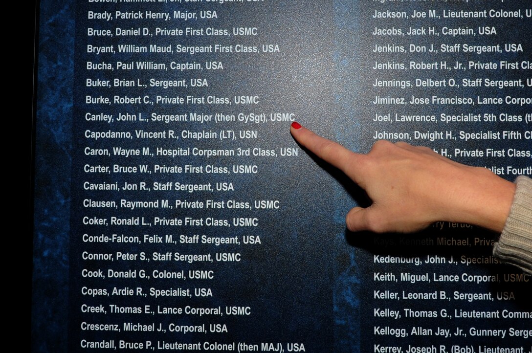 A finger points at a name written in white on list with blue background posted on a wall.