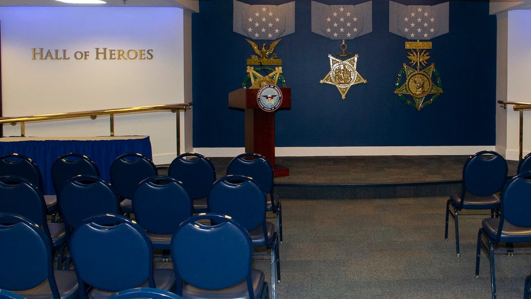 A view of a room with "Hall of Heroes" and three medals on a wall, and audience chairs and a lectern.