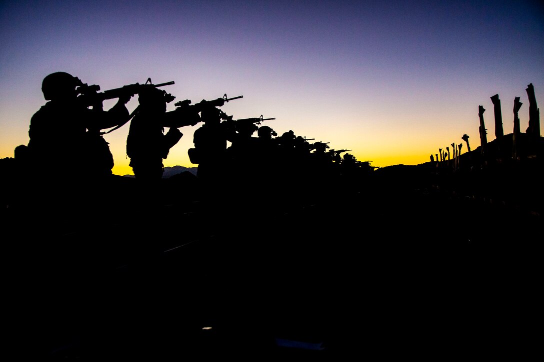 Marines, shown in silhouette, aim weapons in a line against a deep purplish-blue sky.